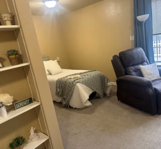 A cozy bedroom with a single bed covered in white bedding and a gray throw blanket. There is a dark recliner chair with a white pillow next to a window with blue curtains. A small bookshelf with decorative items and a sign that reads 'SERENITY' is visible on the left side. The walls are painted beige and the floor is carpeted.