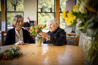Two elderly women sitting at a wooden table in a bright room with large windows, arranging orange flowers in a glass vase. There are additional flowers in the foreground and background, and greenery is visible outside the windows.