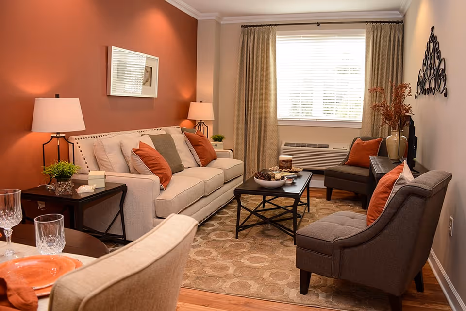 A cozy living room with a beige sofa adorned with orange and gray pillows, two gray armchairs with orange pillows, a black coffee table with decorative items, two side tables with lamps and plants, a large window with beige curtains, and a warm orange accent wall with a framed picture.