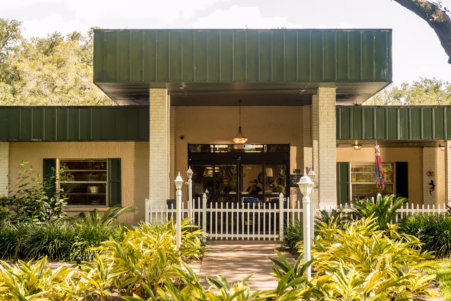 Front exterior view of a single-story building with a green roof and white brick pillars. The entrance has glass doors and is surrounded by a white picket fence with a walkway leading up to it. There are green plants and shrubs in the foreground and trees in the background.