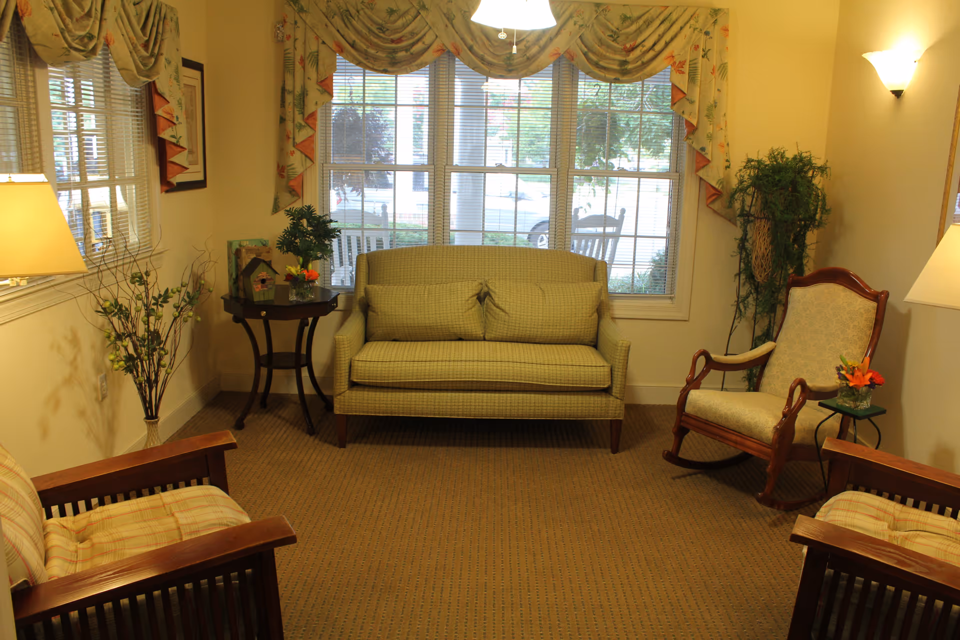 A cozy living room area with a green loveseat centered against a window with floral curtains. Two wooden armchairs with plaid cushions are positioned facing each other, and a wooden rocking chair with a light patterned cushion is on the right. There are two small side tables with plants and flowers, a floor lamp on the left, and a wall sconce on the right, creating a warm and inviting atmosphere.