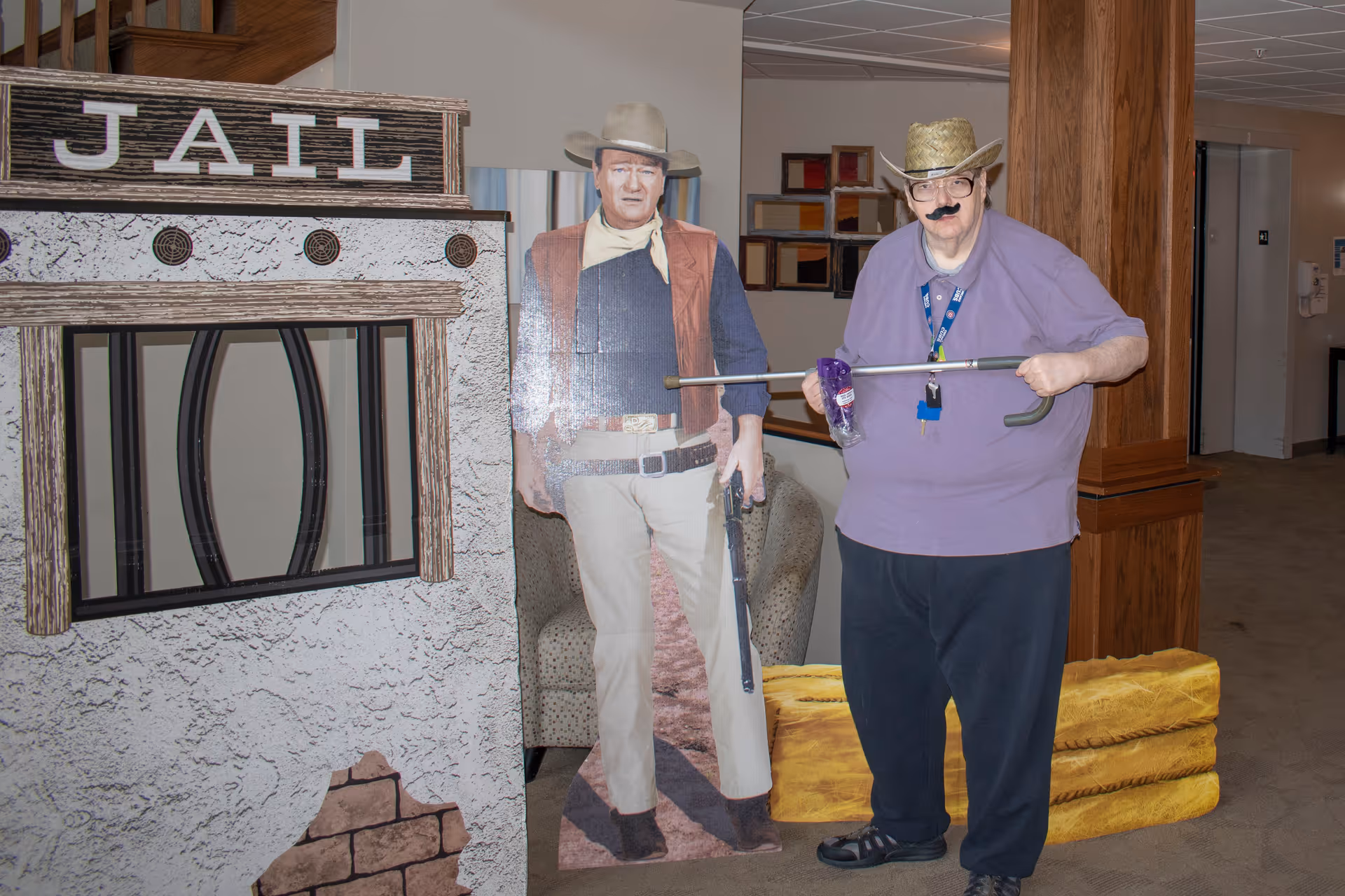 An elderly person wearing a cowboy hat, fake mustache, and purple shirt poses with a toy gun next to a cardboard cutout of a cowboy and a mock jail setup inside a senior living facility.