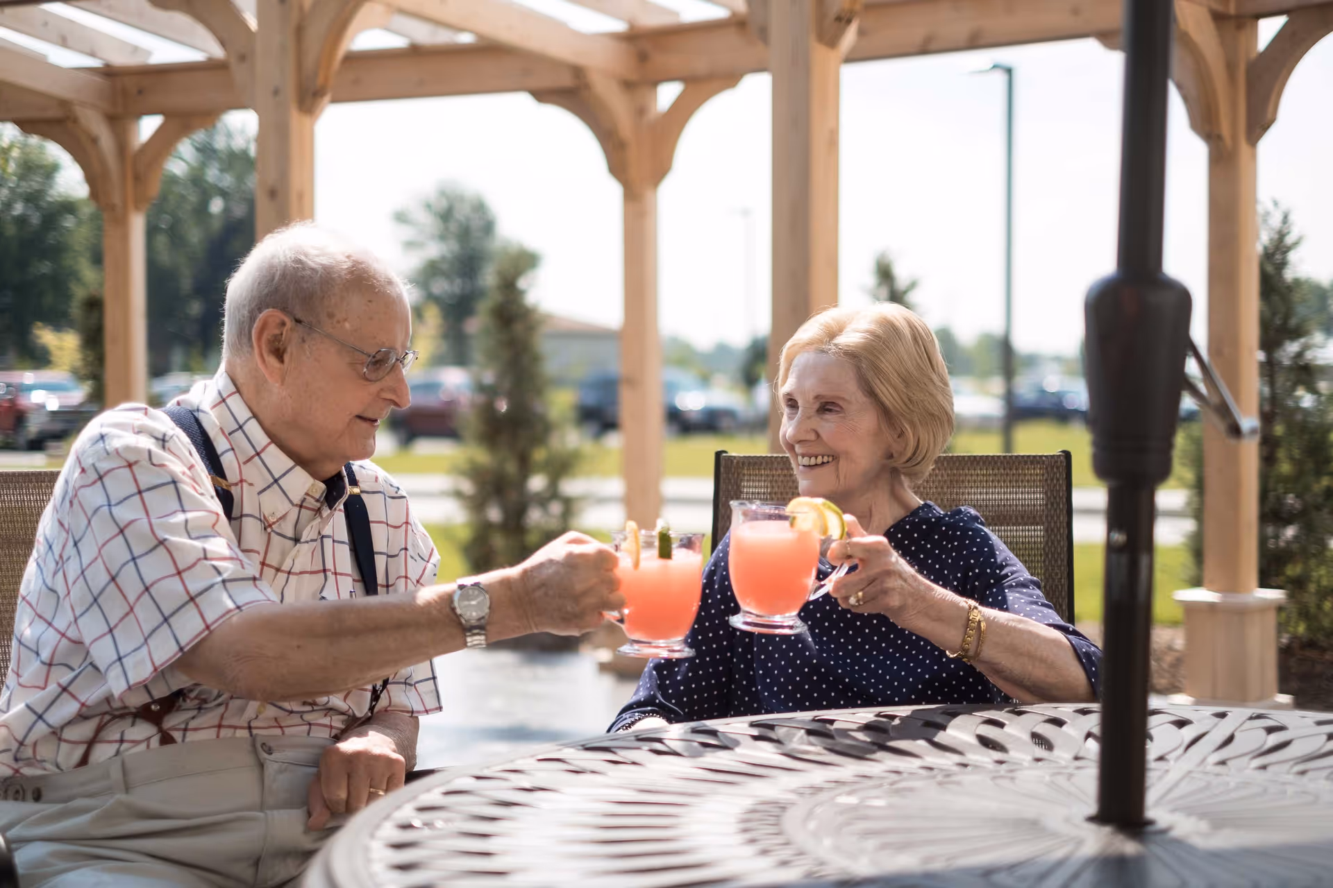 An elderly man and woman sitting outdoors under a wooden pergola, smiling and clinking glasses of pink lemonade garnished with lemon slices.