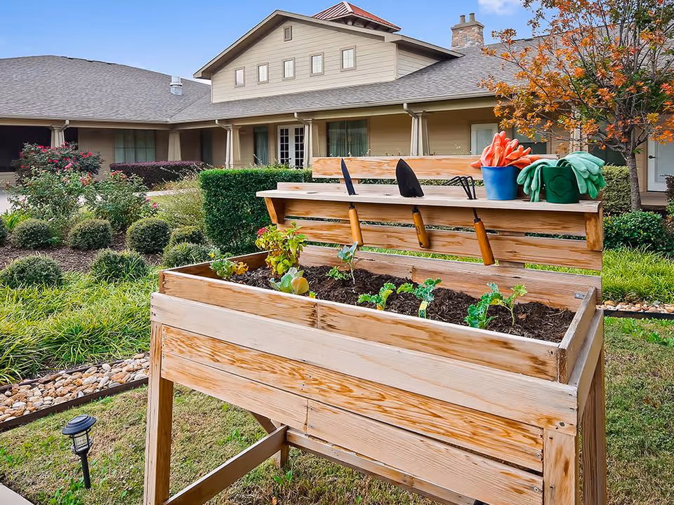 Raised wooden planter with gardening tools and young plants in a landscaped courtyard in front of a residential building.