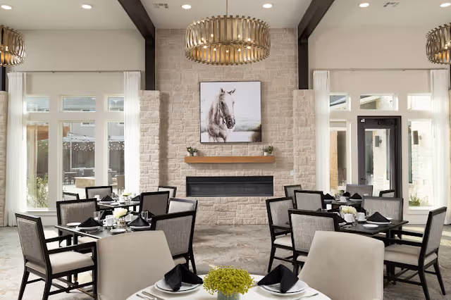 A bright dining room with multiple tables and chairs arranged neatly. Each table is set with white plates, black napkins, and small floral centerpieces. The room features large windows with white curtains, a stone fireplace with a horse painting above it, and a modern chandelier hanging from the ceiling.