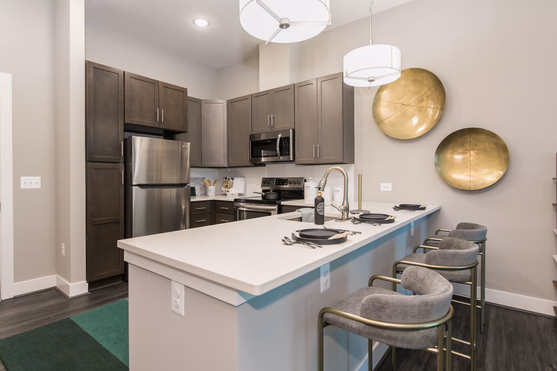 Modern kitchen with dark wood cabinets, stainless steel refrigerator and stove, a white countertop island with a sink, three gray cushioned bar stools with gold frames, and two large decorative gold wall plates. The kitchen is well-lit with two hanging pendant lights.