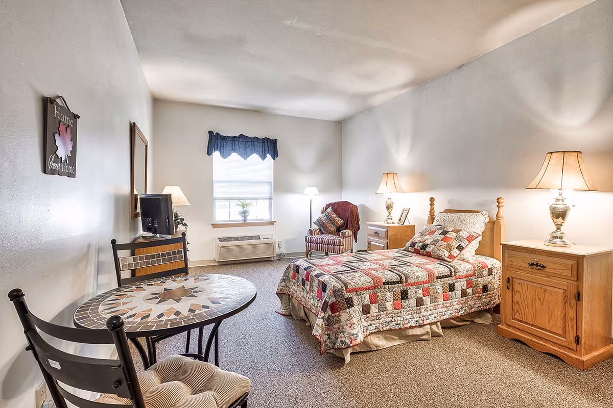 A cozy bedroom in a senior living facility featuring a single bed with a colorful patchwork quilt, two wooden nightstands with lamps, a striped armchair with a throw blanket and pillows, a small round mosaic table with two chairs, a window with a blue valance, and a wall decoration that reads 'Home Sweet Home'.
