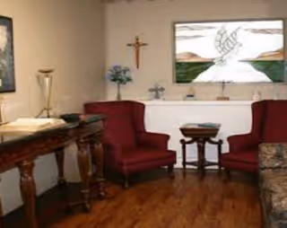 A small, cozy room with two red upholstered armchairs facing each other, a wooden side table between them, and a wooden console table with a lamp and an open book. The room has a wooden floor, a stained glass window with a dove design, a crucifix on the wall, and some decorative items including flowers and a small cross.