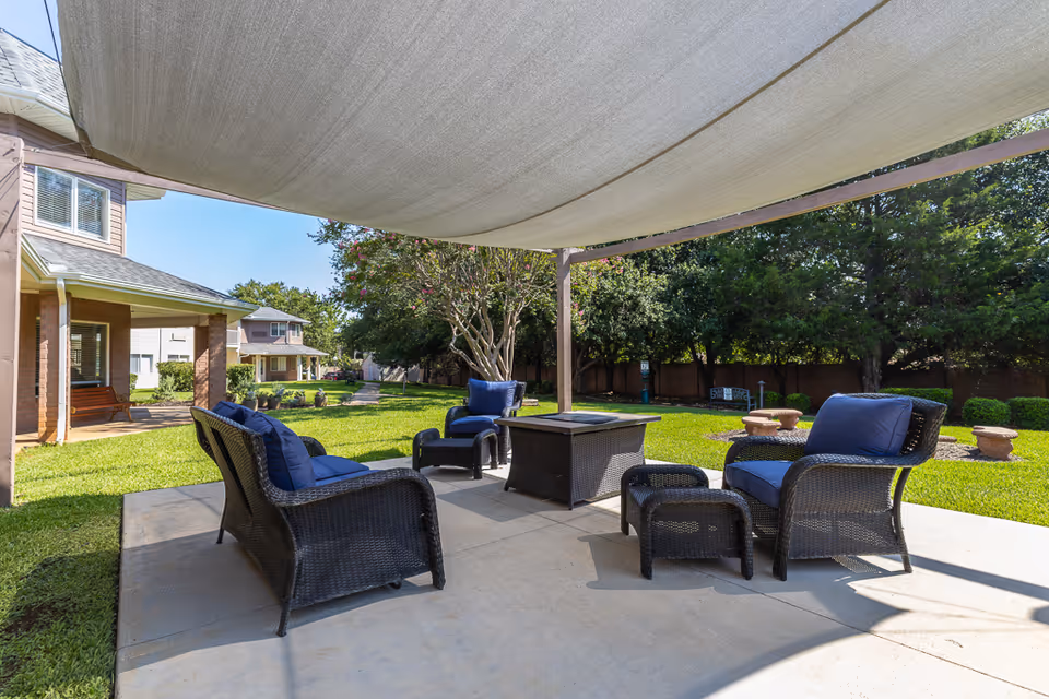 Shaded outdoor patio with wicker chairs and blue cushions arranged around a central table on a lawn near residential buildings.