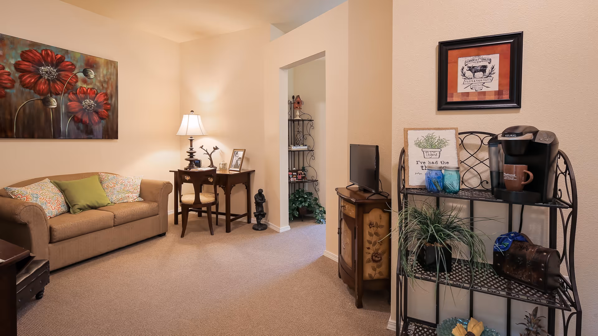 A cozy living room area with a brown sofa adorned with colorful pillows, a wooden desk with a chair and a table lamp, a small TV on a decorative cabinet, and a metal shelf holding a coffee maker, mugs, plants, and decorative items. The walls are beige and decorated with framed artwork.