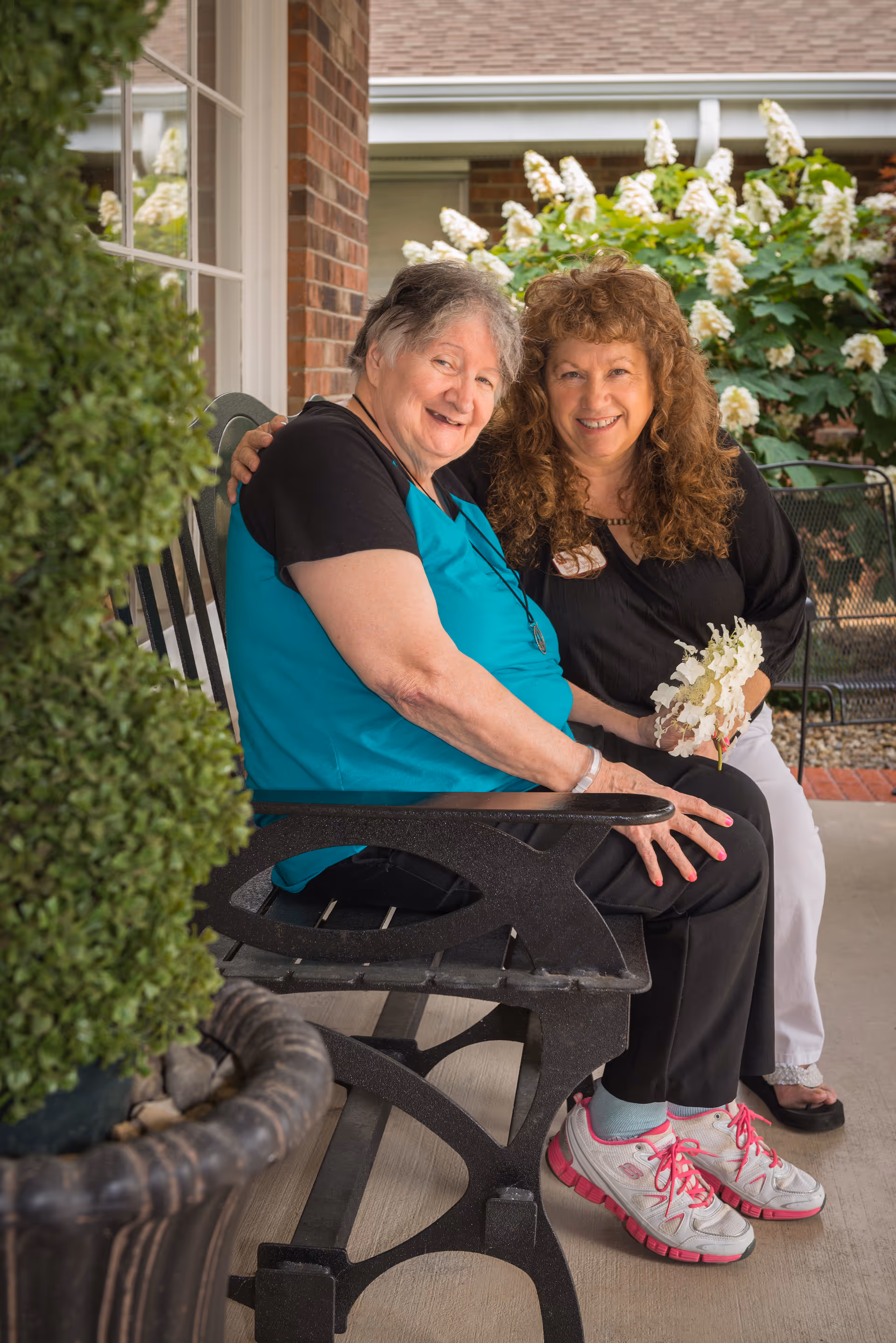 Two women sitting closely together on a black bench outside a brick building, smiling at the camera. One woman is wearing a turquoise and black shirt with black pants and white sneakers with pink accents. The other woman has curly hair, is dressed in black and white, and is holding white flowers. There are green plants and white flowers in the background.
