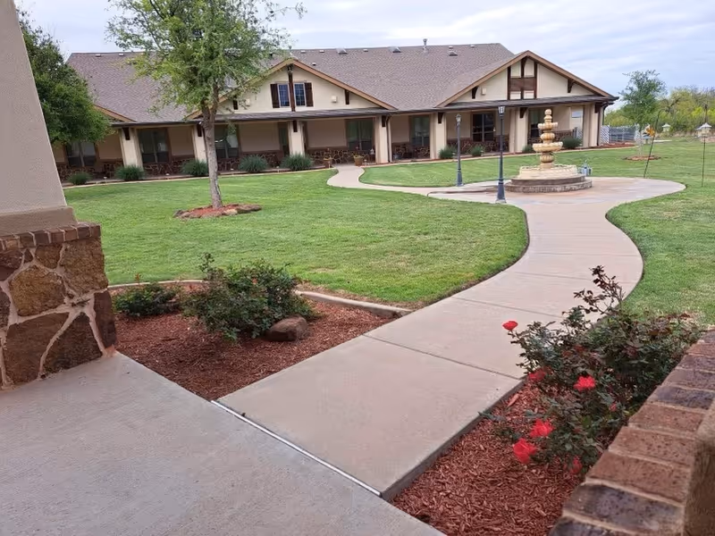 A paved walkway curves through a well-maintained lawn with flower beds and bushes, leading to a multi-unit building with a covered porch and outdoor seating. A decorative fountain and lamp posts are visible along the path under a partly cloudy sky.