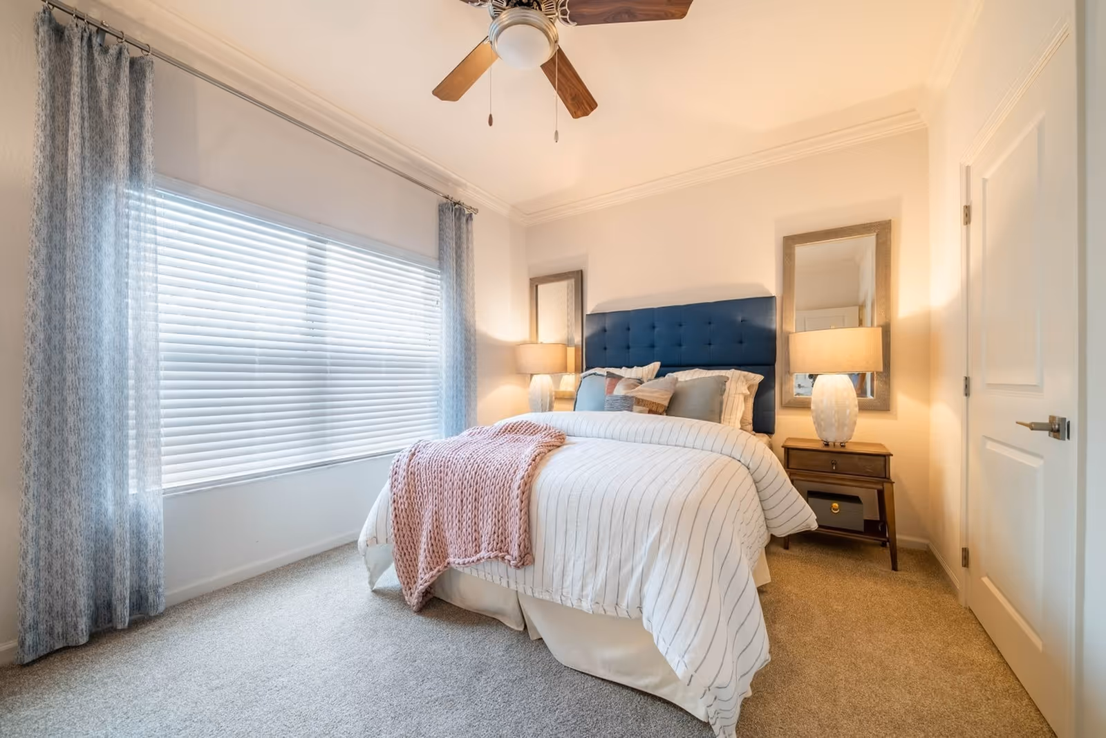A cozy bedroom with a large window covered by blinds and light blue curtains. The bed has a dark blue tufted headboard, white and striped bedding, multiple pillows, and a pink knitted throw blanket. There are two wooden nightstands on either side of the bed, each with a lamp and a rectangular mirror mounted on the wall behind them. The room has beige carpet and a ceiling fan with wooden blades.