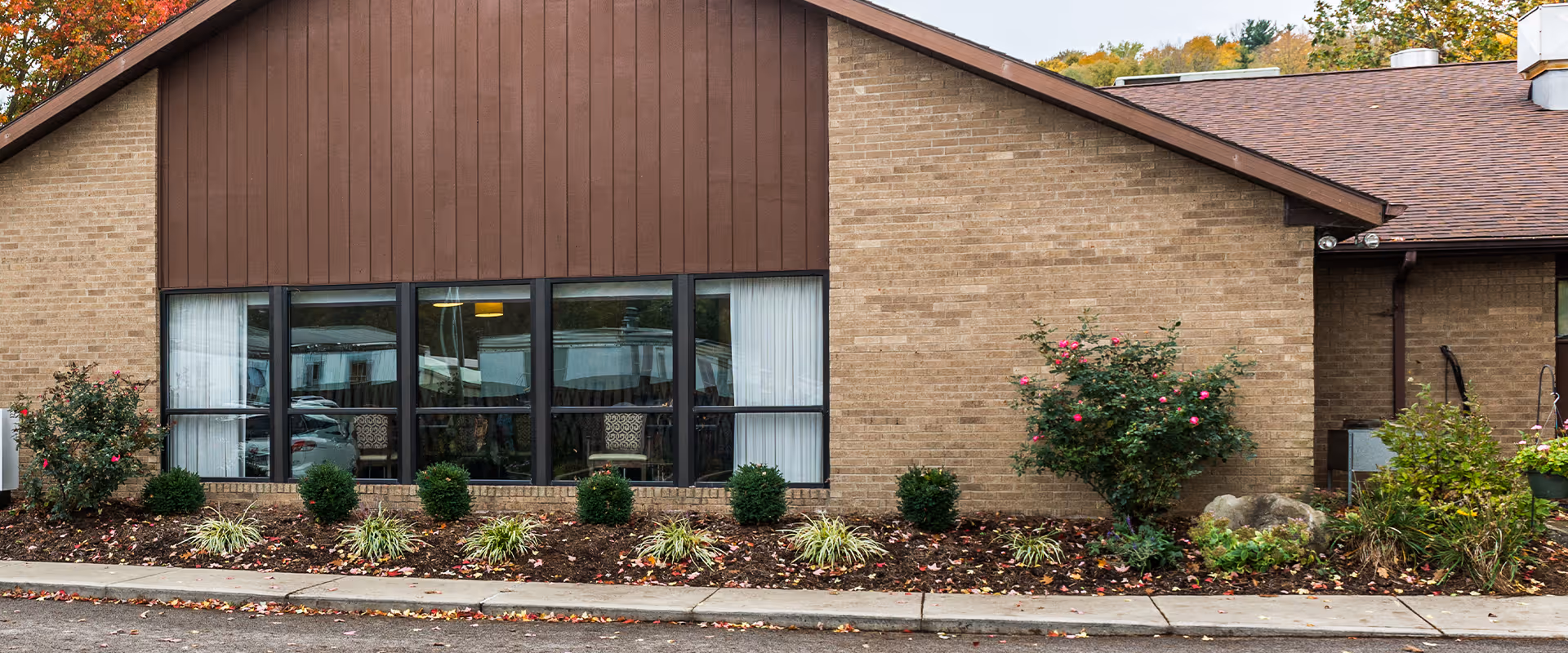 Exterior view of a single-story brick building with a brown roof and large windows. There are shrubs and small plants in front of the building, and some trees with autumn foliage in the background.