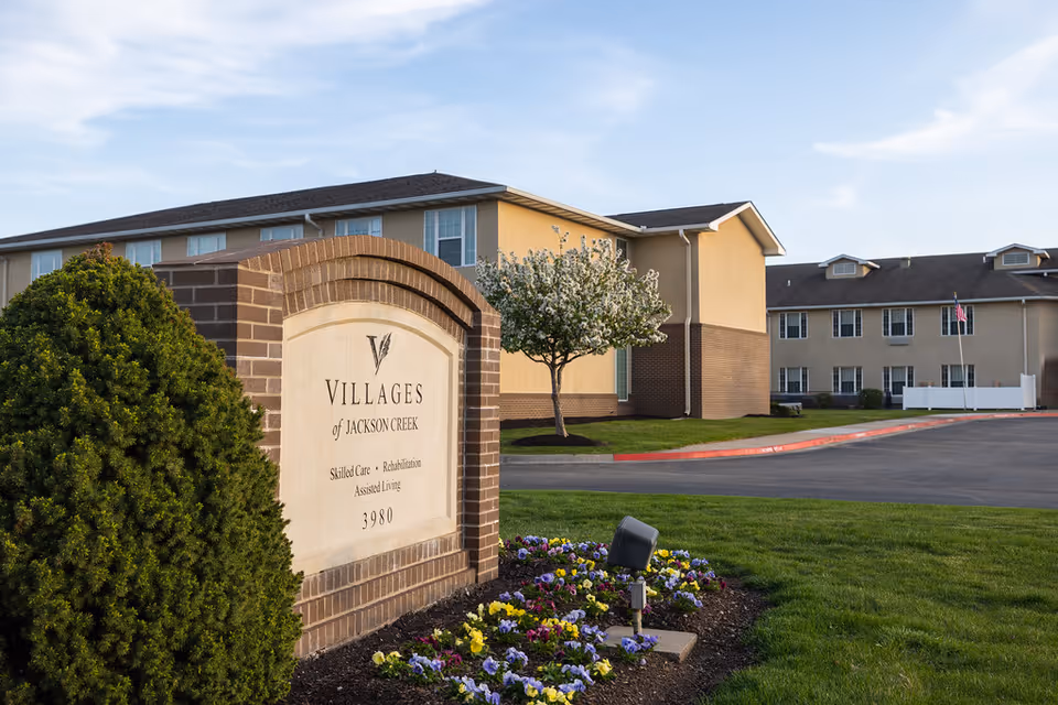 Entrance sign for Villages of Jackson Creek surrounded by flowers and landscaping with the residential facility building in the background.