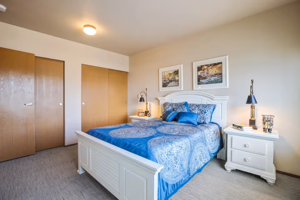 A bedroom with a white wooden bed frame and a blue patterned bedspread. There are two white nightstands on either side of the bed, each with a lamp and decorative items. Two framed pictures hang on the wall above the bed. The room has beige walls, a carpeted floor, and wooden sliding closet doors.