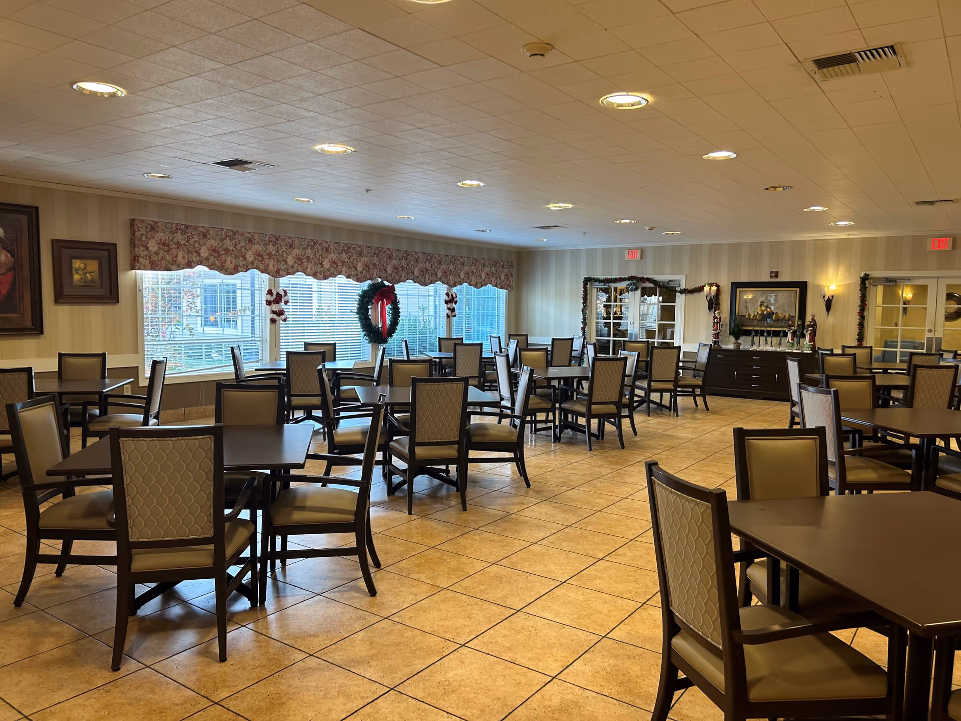 A spacious dining room with multiple dark wood tables and cushioned chairs arranged neatly. Large windows with floral valances allow natural light to brighten the room. The walls are decorated with framed paintings and holiday wreaths, and there is a sideboard with decorative items along one wall. The floor is tiled, and the ceiling has recessed lighting.