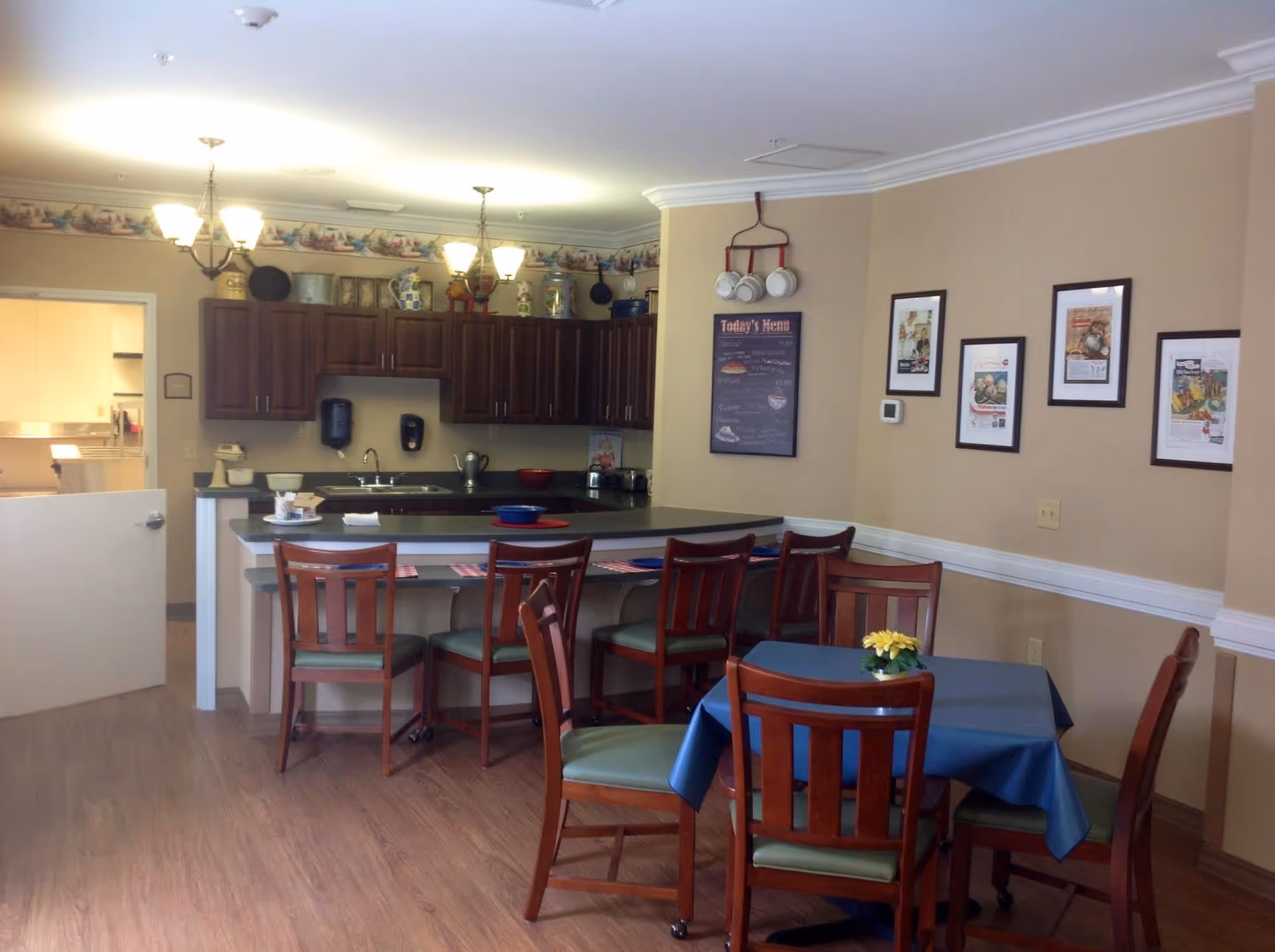 A dining area with a kitchen counter and several wooden chairs. The kitchen has dark wood cabinets, a sink, and various kitchen items on the counter and shelves. There is a small table with a blue tablecloth and a yellow flower centerpiece. The walls are beige with framed pictures and a menu board hanging. The floor is wooden, and the room is well-lit with ceiling lights.