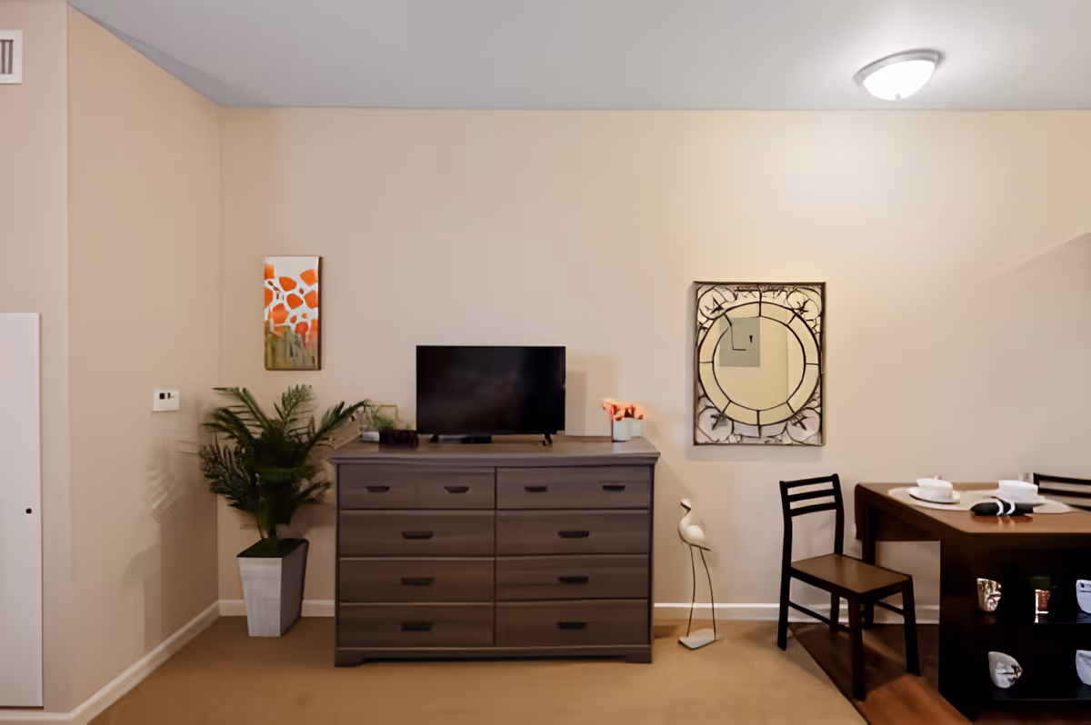 Interior view of a room with beige walls featuring a dark wooden dresser with a flat-screen TV on top, a potted green plant to the left, a decorative wall mirror and artwork above the dresser, and a dark wooden dining table with two chairs set with plates and napkins on the right.