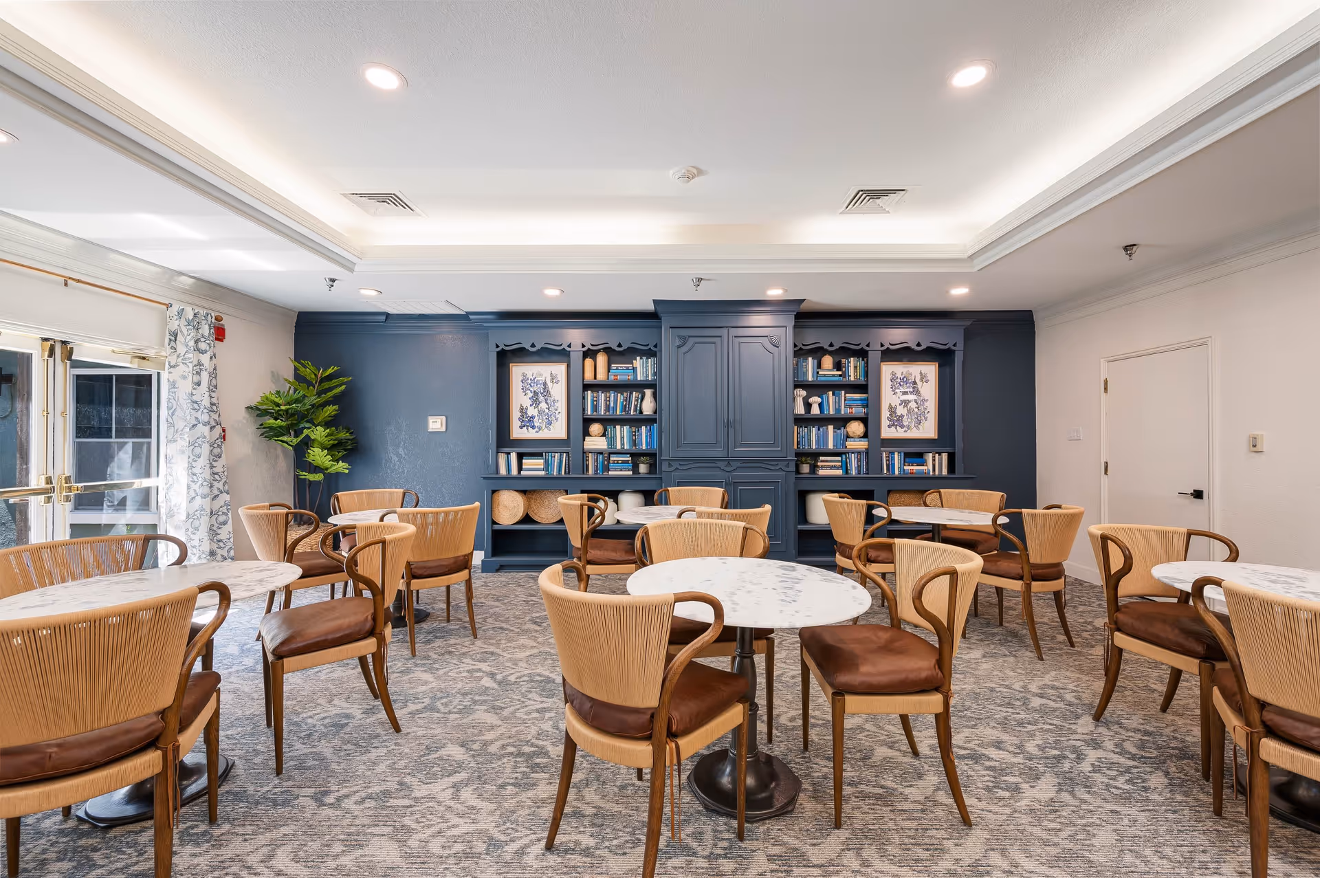 A bright and spacious dining room with multiple round tables and wooden chairs with brown cushions. The back wall features a large built-in blue bookshelf filled with books and decorative items, flanked by two framed floral artworks. There is a large window with floral curtains on the left side and a potted plant near the bookshelf. The ceiling has recessed lighting and crown molding.