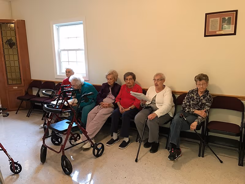 Six elderly women sitting on chairs along a wall in a room with a window and a wooden door. Two walkers are positioned in front of them. One woman is holding papers and appears to be speaking or reading.