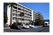 Exterior front view of a multi-story building with a parking lot, several cars, palm trees, and a blue sky.
