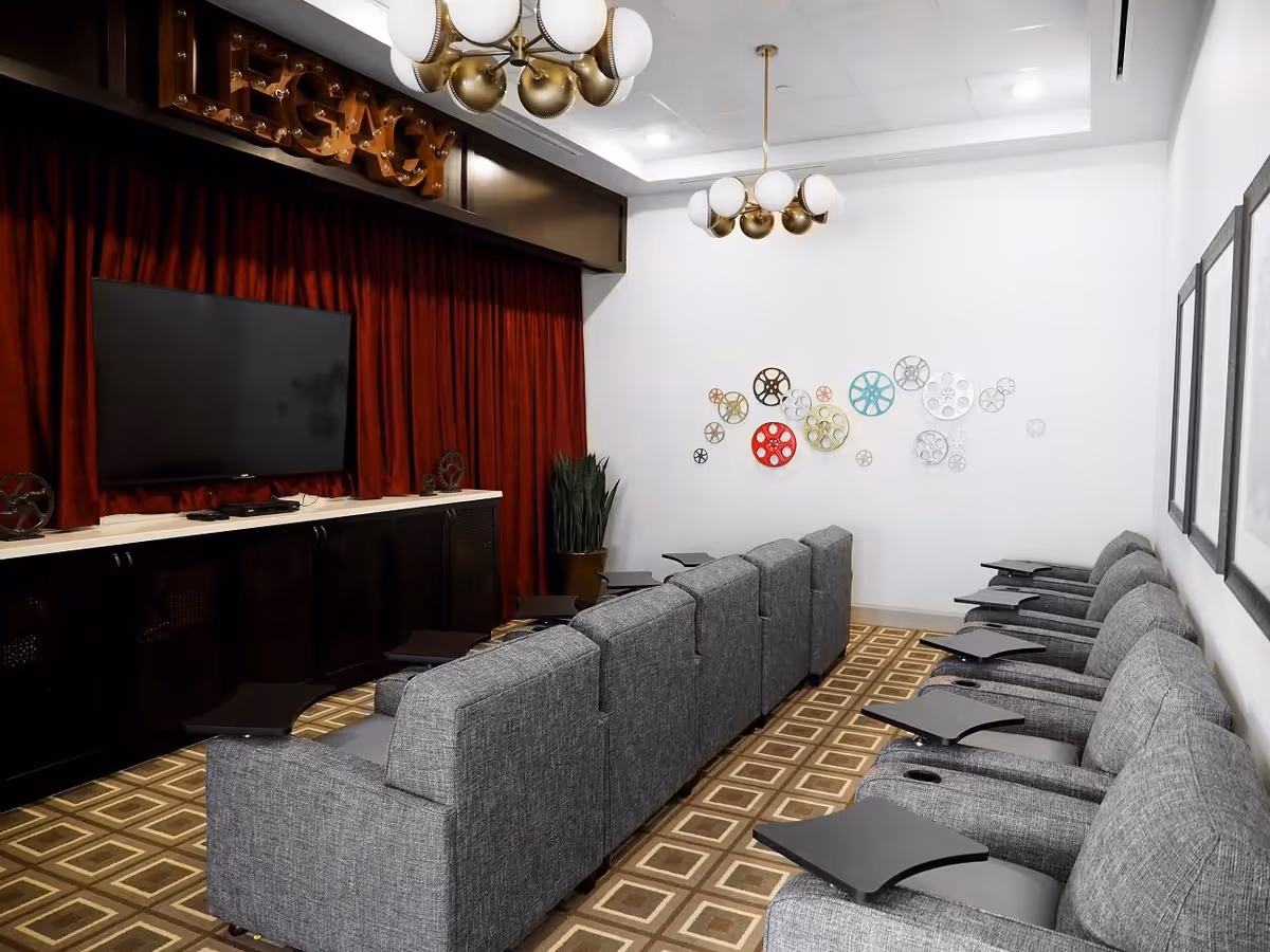 A small theater room with two rows of gray upholstered reclining chairs, each equipped with a black tray table. The room features a large flat-screen TV mounted on a wall with red curtains behind it. The walls are white with decorative film reel wall art and framed pictures. The ceiling has modern brass and white globe light fixtures, and the floor is covered with a patterned carpet.