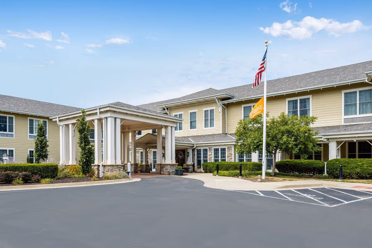 Exterior view of Sunrise of Overland Park senior living facility showing a two-story building with beige siding, multiple windows, a covered entrance with white columns, an American flag, and a Sunrise flag on flagpoles, surrounded by neatly trimmed bushes and a paved driveway.