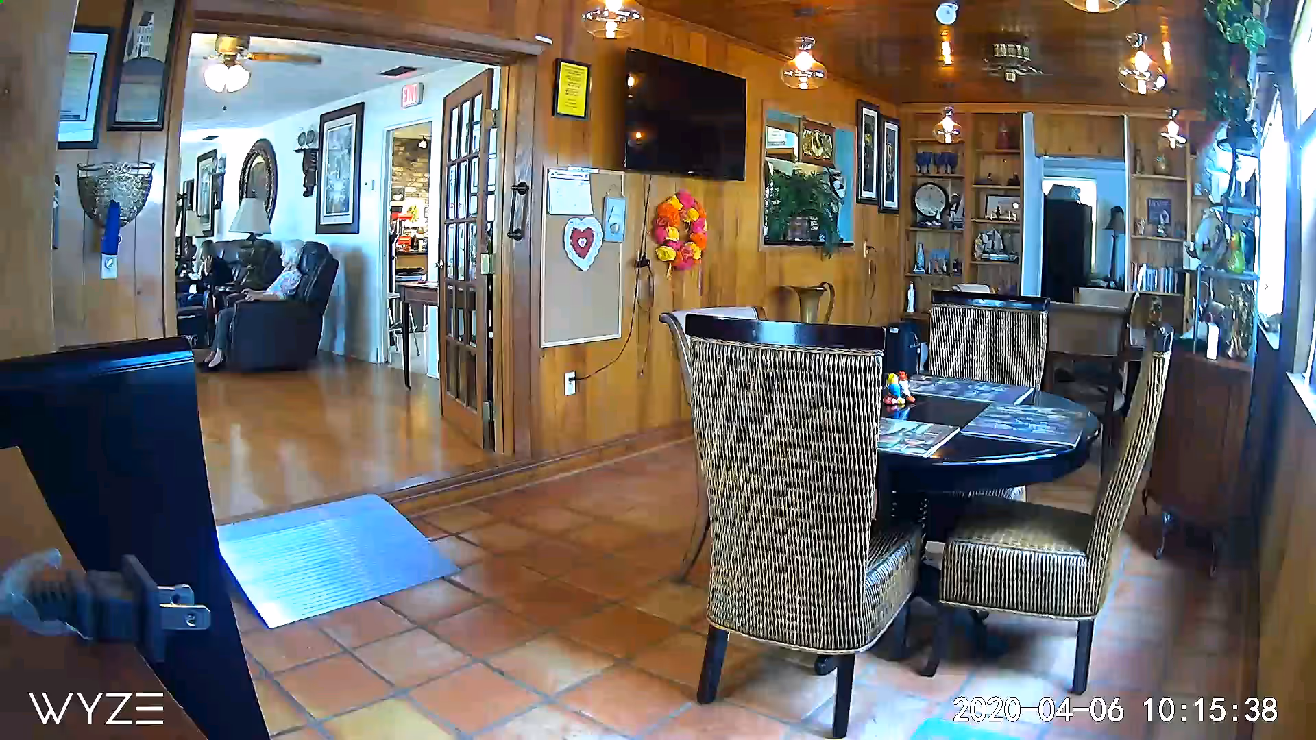 Interior view of a senior living facility showing a dining area with a round table and four wicker chairs. The room has wood-paneled walls, a mounted TV, decorative items, and a bulletin board. Through an open doorway, a living room area with recliner chairs and framed pictures on the walls is visible. The floor is tiled in the dining area and wooden in the living room.