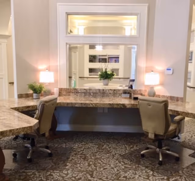 Interior view of a reception or common area with a large marble countertop desk, two beige office chairs on wheels, two table lamps with white shades, a potted plant, and a large mirror on the wall reflecting another room with a plant and windows.