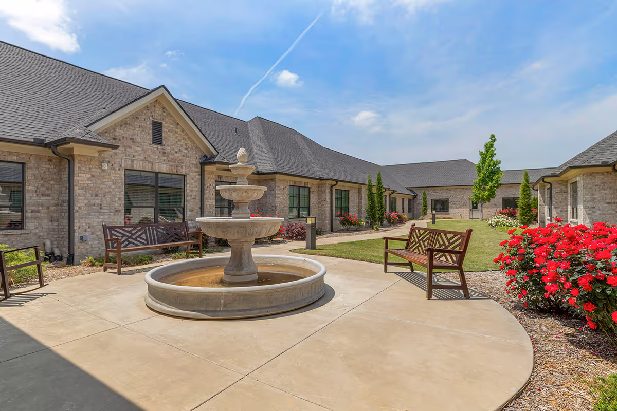Outdoor courtyard area at Bellarose Senior Living featuring a central stone fountain surrounded by benches, with brick buildings, green grass, trees, and flowering bushes under a partly cloudy blue sky.