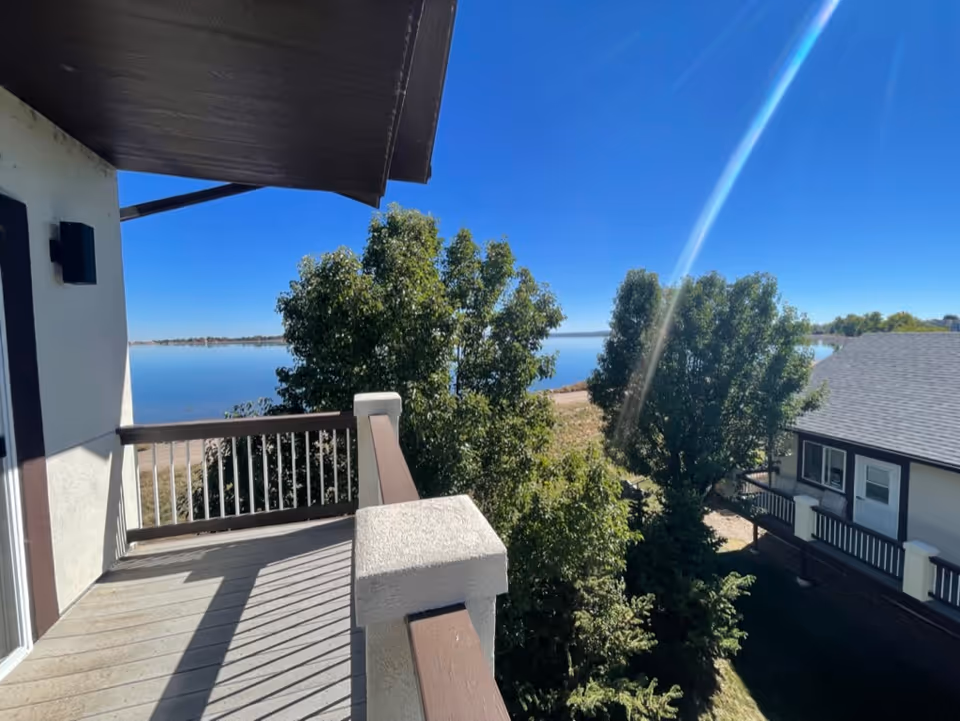 View from a balcony overlooking trees and a large body of water under a clear blue sky. Part of a neighboring building with a gray roof and white walls is visible on the right side.