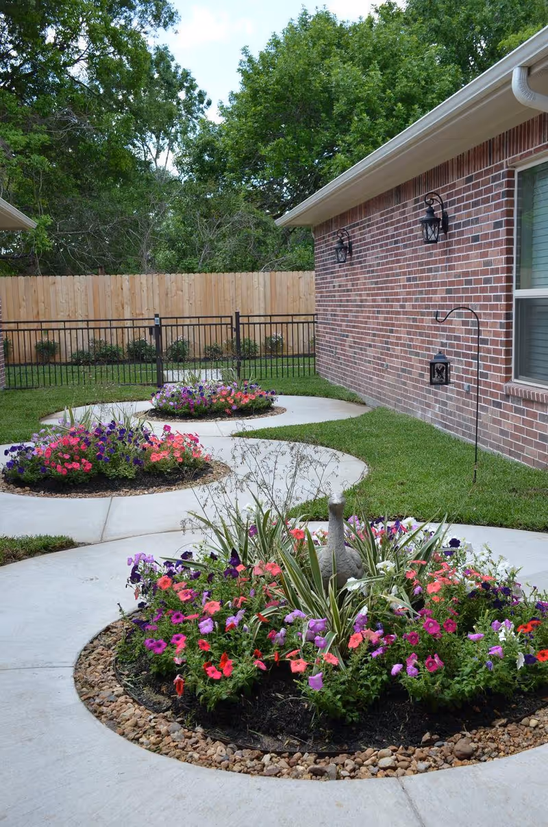 A landscaped outdoor garden area with circular flower beds filled with colorful flowers and greenery. The garden is bordered by a concrete walkway and a brick building wall with outdoor lantern-style lights. There is a wooden fence and trees in the background.