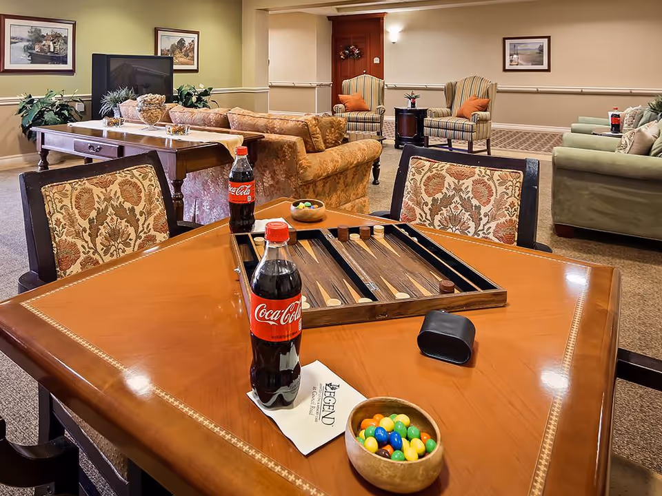 A cozy common area in a senior living facility featuring a wooden table with a backgammon board, two bottles of Coca-Cola, a bowl of colorful candy, and a napkin with the Legend At Council Road logo. The background shows comfortable upholstered chairs, sofas, framed artwork on the walls, and plants, creating a warm and inviting atmosphere.