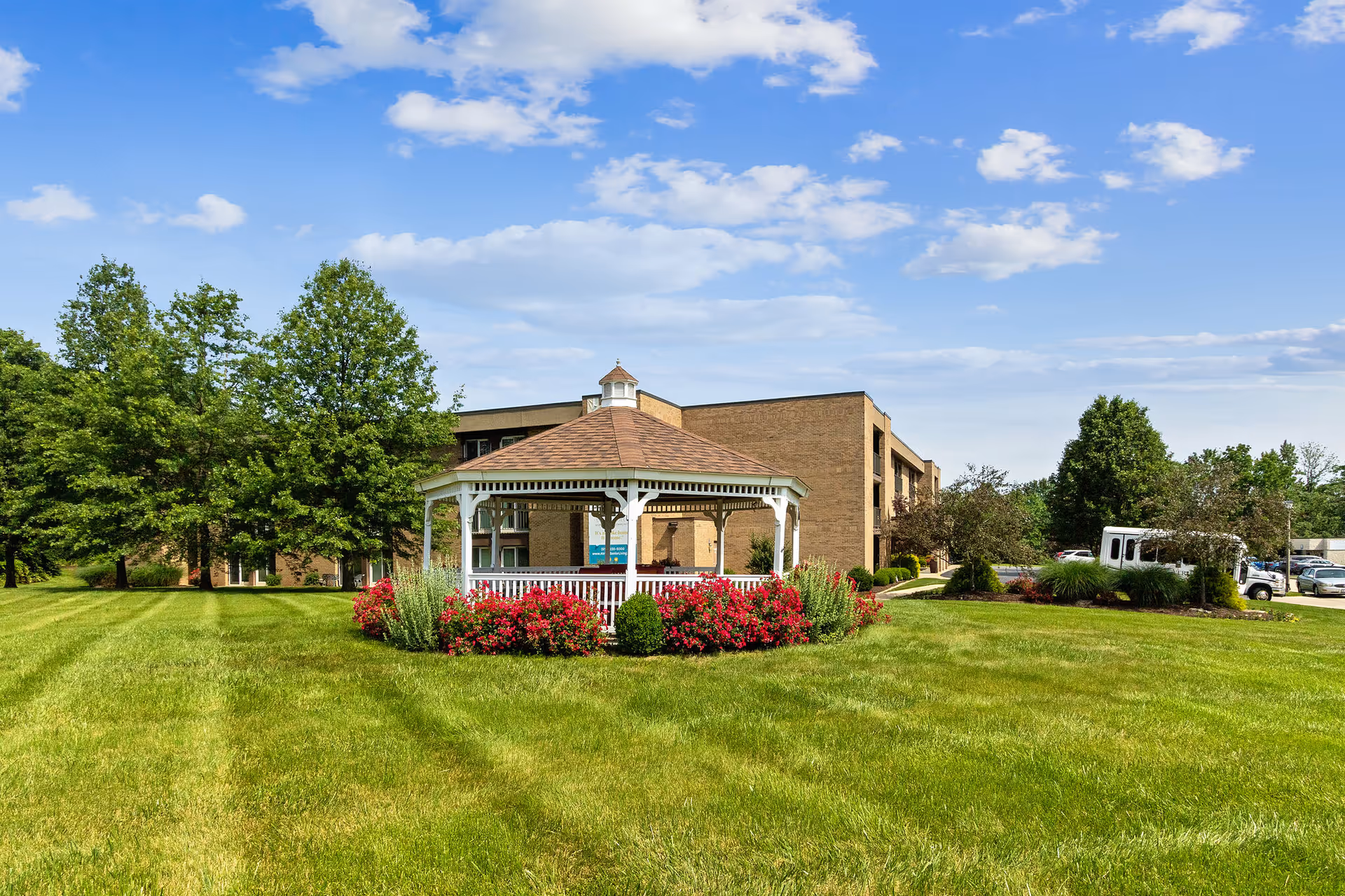 A white gazebo with a brown shingled roof surrounded by red flowers and green shrubs sits on a well-maintained grassy lawn. Behind the gazebo is a three-story brick building with balconies, and there are several trees and a parking area with vehicles in the background under a partly cloudy blue sky.