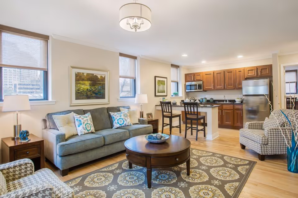 A bright and modern living room and kitchen area in an apartment. The living room features a gray sofa with decorative pillows, two patterned armchairs, a round wooden coffee table on a patterned rug, and two side tables with lamps. The kitchen has wooden cabinets, a stainless steel refrigerator, a microwave, and a small breakfast bar with two chairs. Large windows with blinds allow natural light to fill the space.
