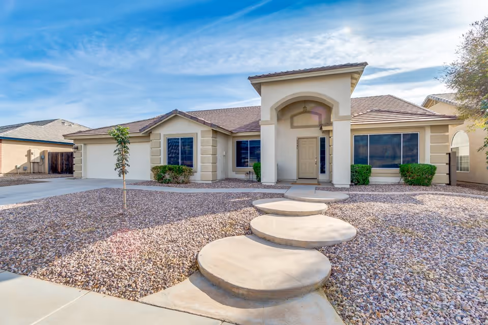 Front exterior of a single-story beige house with circular concrete stepping stones leading across a gravel yard to the covered entrance and attached garage.