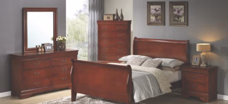 A bedroom with wooden furniture including a bed with pillows and a blanket, a dresser with a mirror, a tall chest of drawers, and a nightstand with a lamp and framed photo. Two framed floral pictures hang on the wall above the bed.
