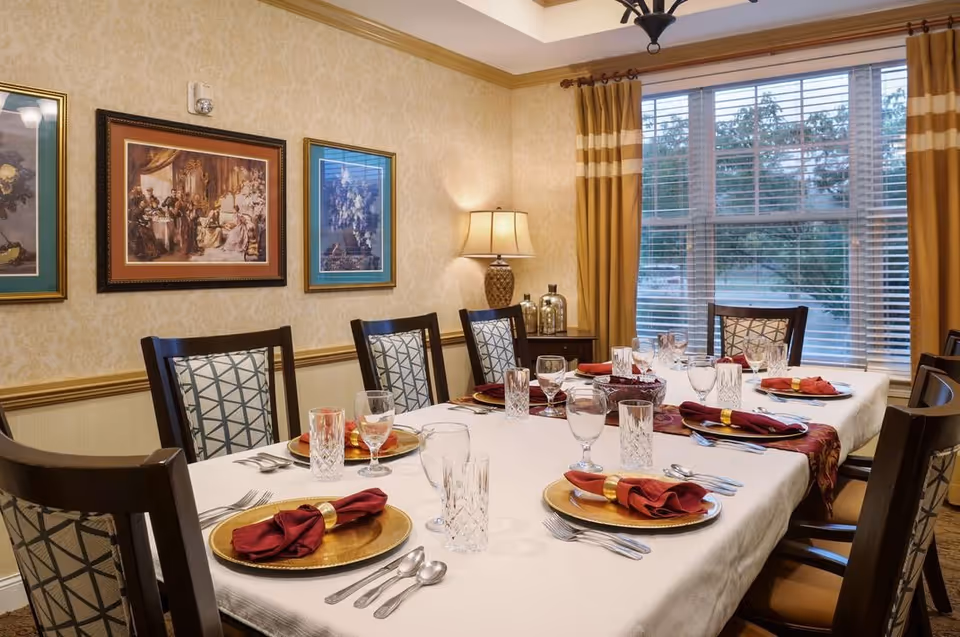 A neatly set dining table with place settings, glassware and folded napkins in a warmly decorated room with framed art and a large window.