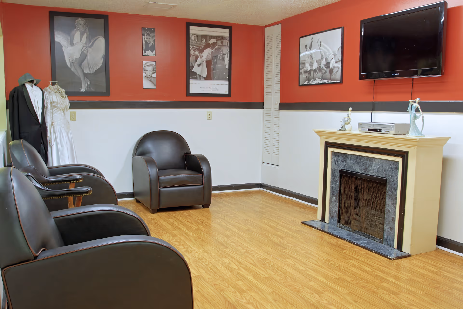 A cozy living room with three black leather armchairs arranged facing a fireplace with a TV mounted above it. The walls are painted red on the upper half and white on the lower half, decorated with black and white framed photographs. Two mannequins dressed in vintage clothing stand in the corner.