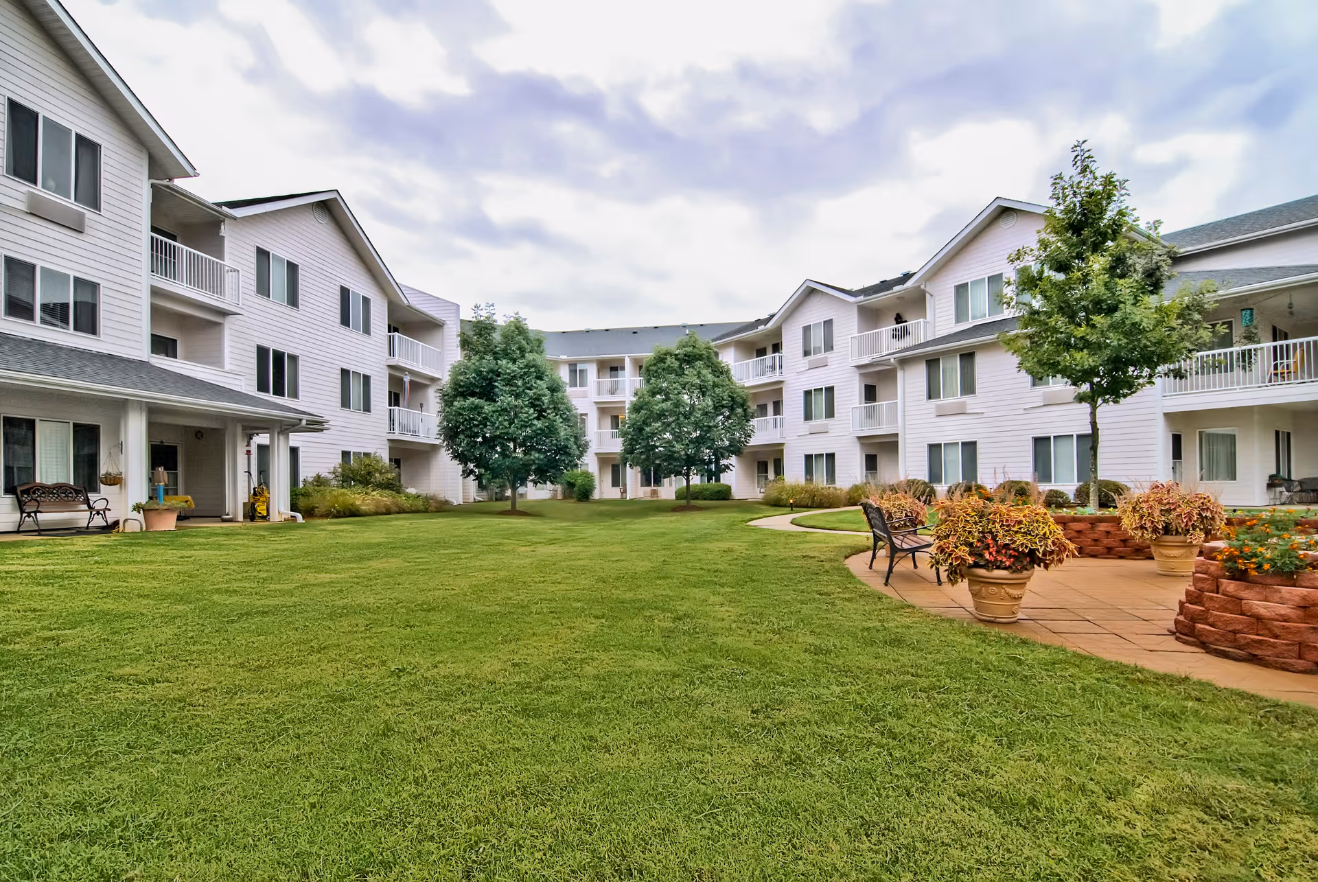 A spacious outdoor courtyard area of a senior living facility with well-maintained green grass, several trees, benches, potted plants, and a paved walkway. Surrounding the courtyard are three-story white residential buildings with balconies and windows.