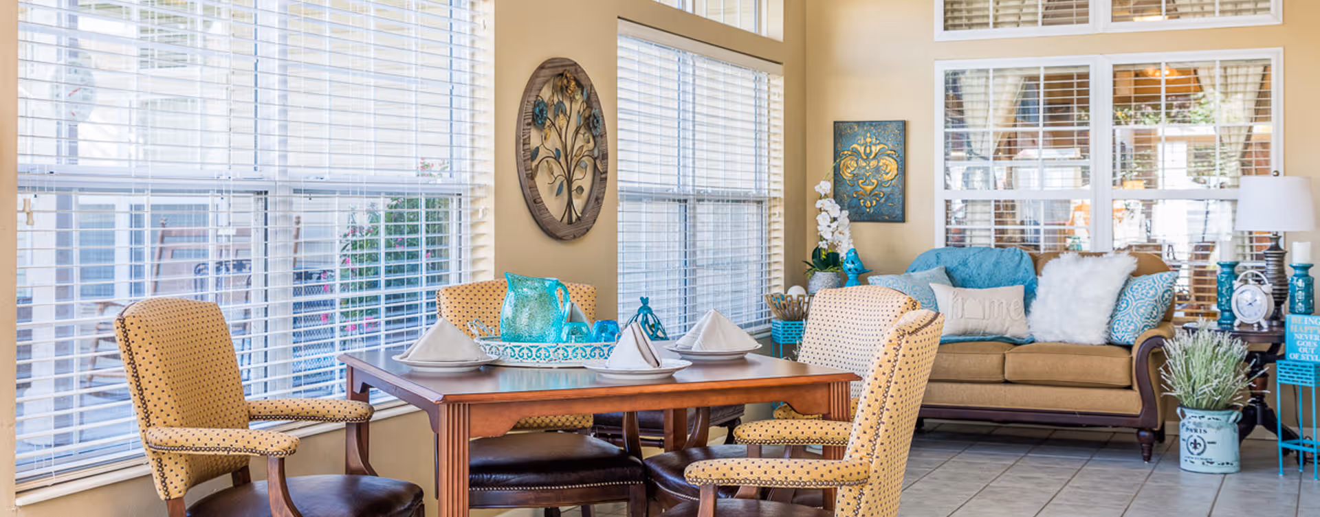 A bright and cozy living area with a wooden dining table set for four, featuring yellow upholstered chairs with nailhead trim. Large windows with white blinds let in natural light. In the background, there is a beige sofa adorned with blue and white pillows, a side table with a lamp, and decorative wall art. The room has a tiled floor and a warm, inviting atmosphere.