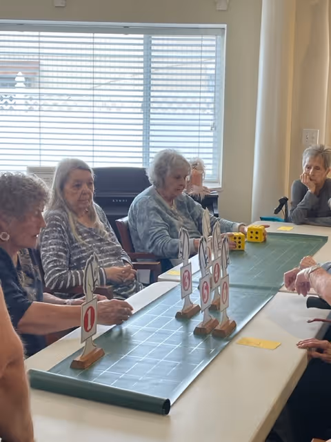A group of elderly women sitting around a table playing a tabletop game with numbered wooden stands and large dice in a well-lit room with a window covered by blinds.