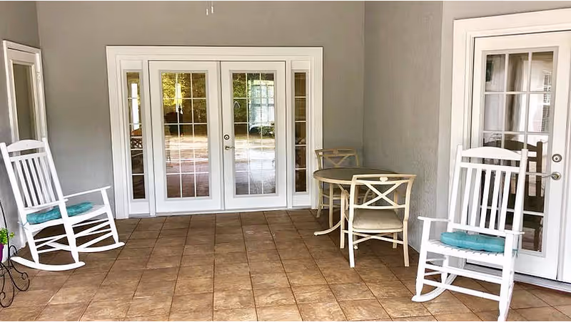 A covered patio area with tiled floor featuring two white rocking chairs with blue cushions, a small round table with two chairs, and white-framed glass doors leading inside.