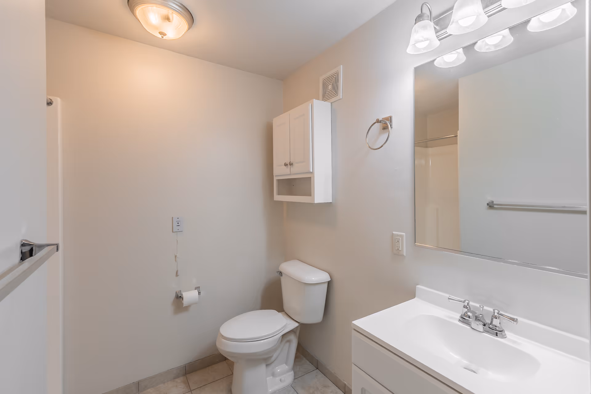 A clean bathroom with beige walls and tiled floor featuring a white toilet, a white vanity with a sink and faucet, a large mirror above the vanity, a wall-mounted cabinet above the toilet, a towel ring, and a ceiling light fixture.