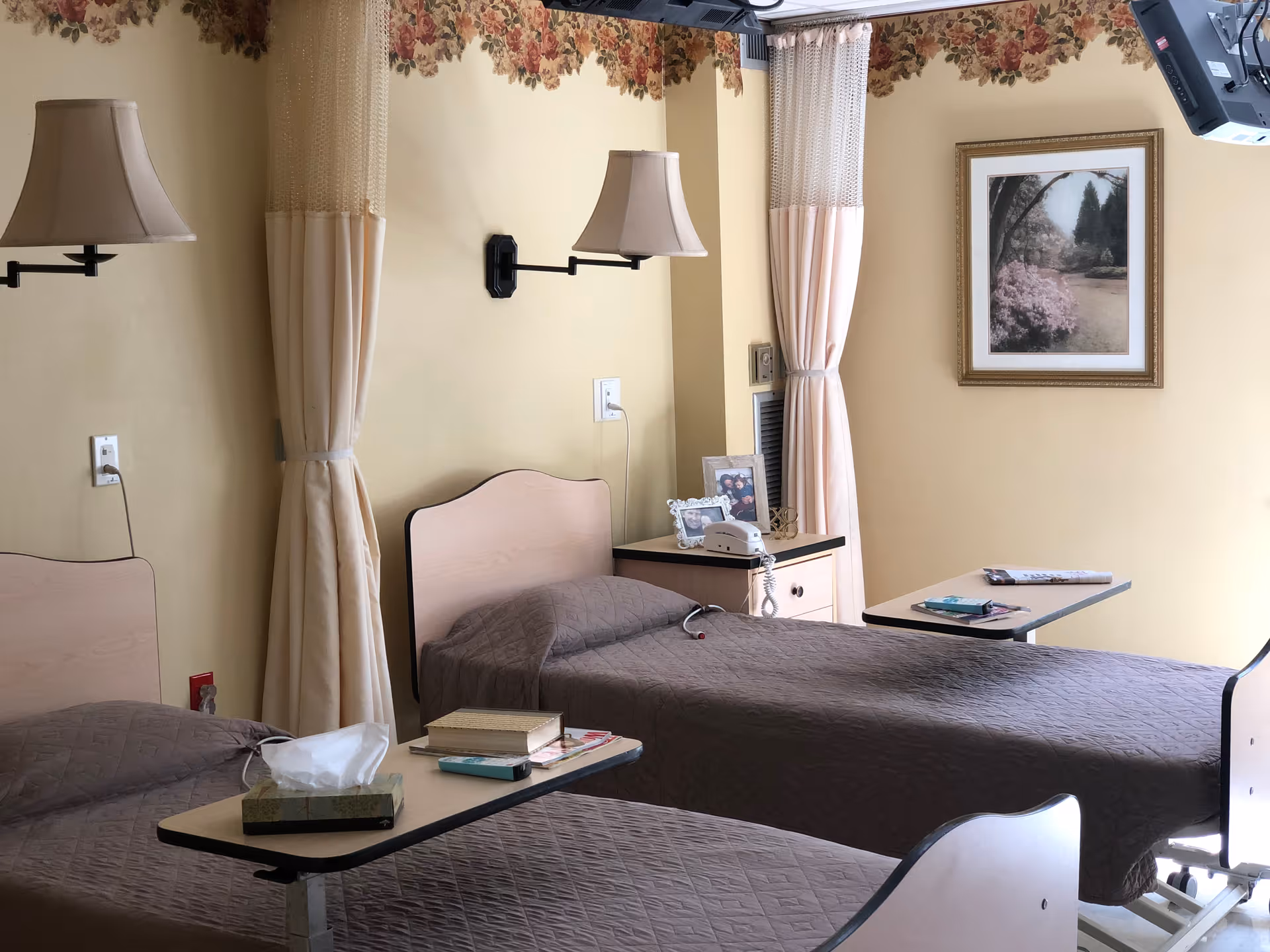 A room with two single beds covered with brown quilts, each with an overbed table holding books, tissues, and other items. The room has beige walls with floral wallpaper border near the ceiling, two wall-mounted lamps, curtains, a nightstand with a telephone and framed photos, and a framed picture on the wall.