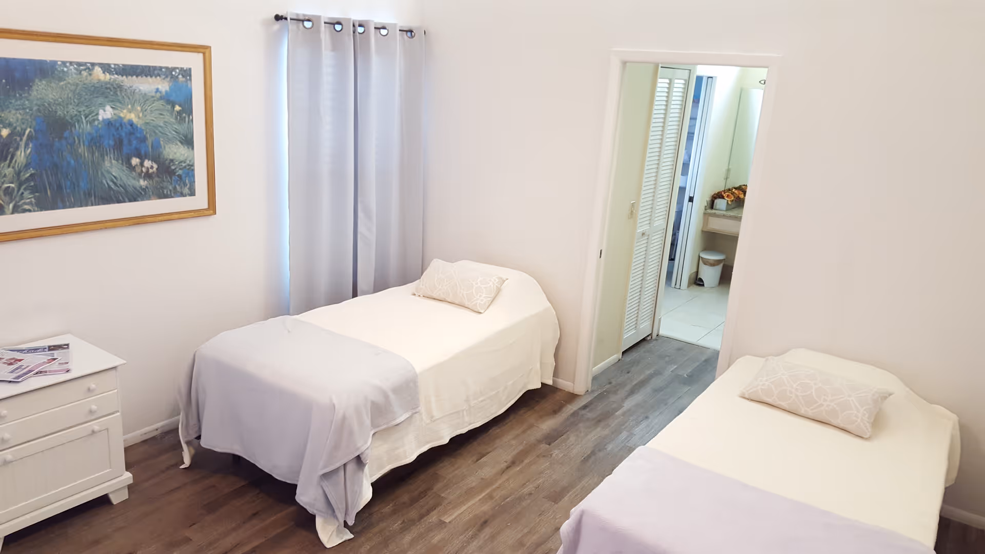 A bright bedroom with two single beds, each with white bedding and a light purple throw blanket. There is a small white nightstand with magazines on it, a window with light gray curtains, and a framed painting on the wall. A doorway leads to a bathroom area with a sink and a trash bin visible.