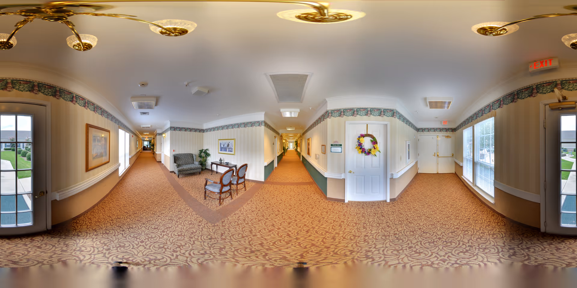 A wide-angle view of a well-lit hallway in a senior living facility with patterned carpet and wallpaper. The hallway features seating areas with chairs and a small table, framed artwork on the walls, and large windows letting in natural light. There are exit doors on both ends of the hallway, and a door decorated with a colorful wreath.
