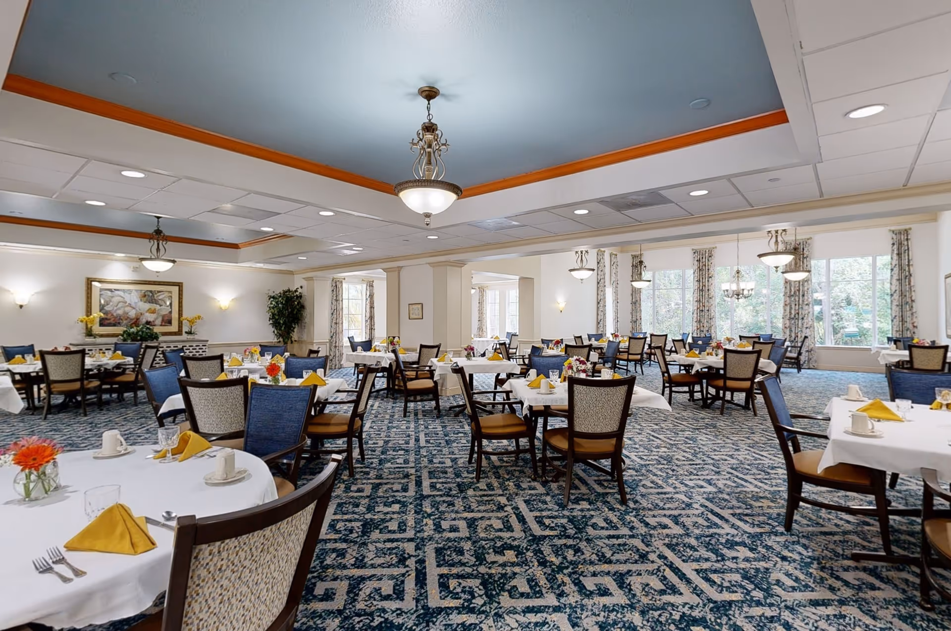 A spacious dining room with multiple round tables covered with white tablecloths, each set with yellow napkins, cups, and small flower arrangements. The room has patterned blue carpet, large windows with floral curtains, and elegant ceiling lights. The walls are decorated with framed artwork and plants.