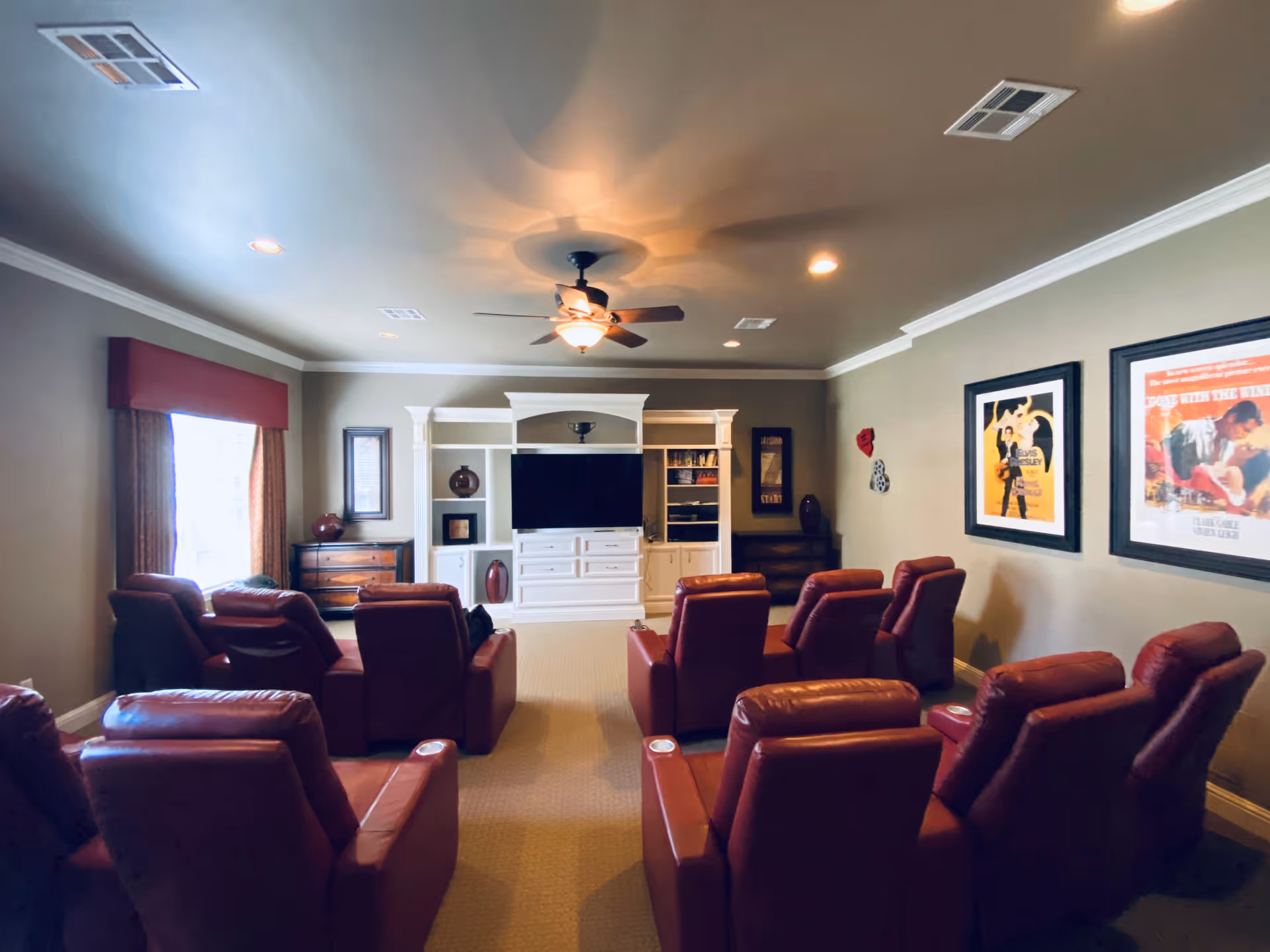 A cozy living room with multiple red leather recliners arranged in rows facing a large flat-screen TV mounted on a white entertainment center. The room has beige walls, a ceiling fan with a light, framed movie posters on one wall, and windows with curtains on the left side.